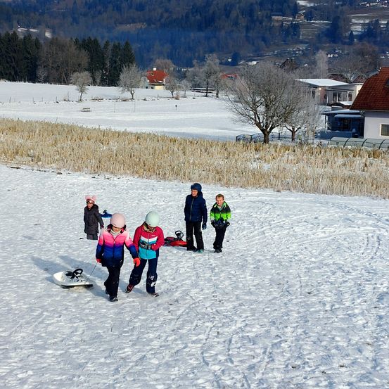 Mehrere Kinder gehen durch ein verschneites Feld. Sie sind warm in Winterkleidung gekleidet und einige halten Schlitten. Ein Haus mit rotem Dach ist im Hintergrund.