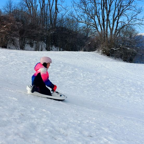 Ein kleines Kind schlitten auf einem schneebedeckten Hügel hinunter, mit kahlen Bäumen im Hintergrund. Sie trägt einen Helm und Handschuhe.
