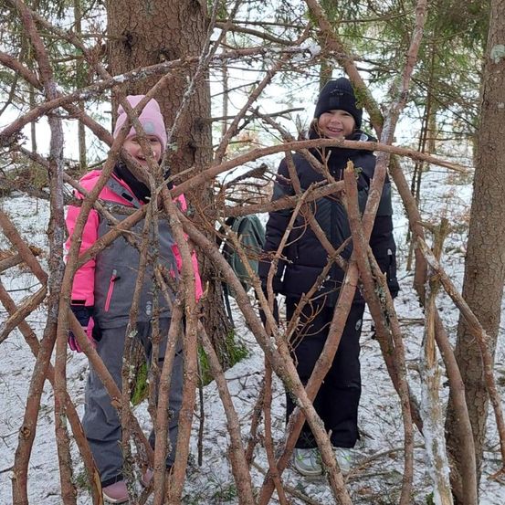 Zwei Kinder, eines in einer pinken Jacke und das andere in einer schwarzen Jacke, stehen in einem verschneiten Wald. Sie lächeln und scheinen für ein Foto zu posieren.