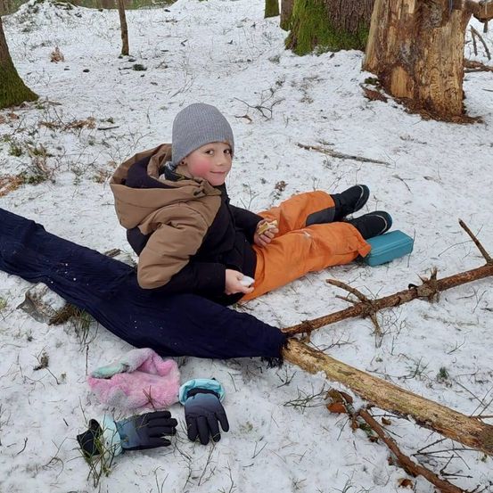 Ein Kind sitzt auf einem Ast im Schnee und trägt eine warme Winterjacke und Handschuhe. Es lächelt und isst einen Snack.
