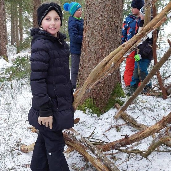 Eine Gruppe von Kindern steht in einem verschneiten Wald, einige klettern auf Äste, einer lächelt im Vordergrund, andere stehen hinter einem Baum.
