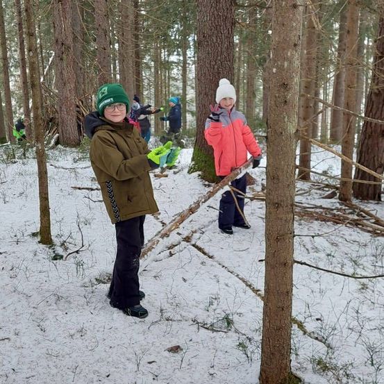 Zwei Kinder in Wintermänteln halten Stöcke in einem verschneiten Wald, mit anderen Kindern und Erwachsenen im Hintergrund.