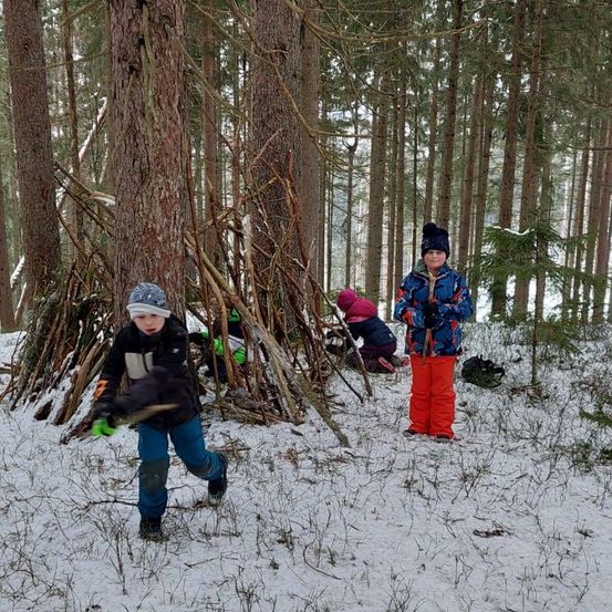 Mehrere Kinder spielen in einem verschneiten Wald. Ein Kind läuft, und zwei andere bauen eine Struktur mit Stöcken.