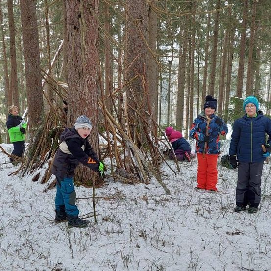 Kinder sind in einem verschneiten Wald, eines sammelt Stöcke. Ein Kind spielt in der Nähe einer Baumstruktur. Hohe Bäume umgeben das Gebiet.