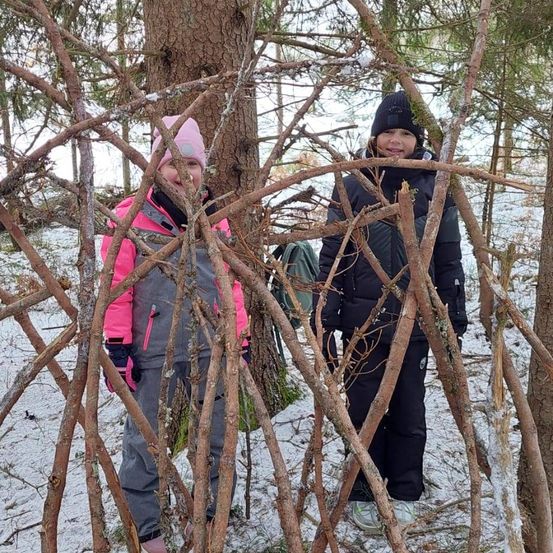 Zwei junge Mädchen stehen hinter einer Wand aus Zweigen in einem verschneiten Wald, beide tragen Winterkleidung.