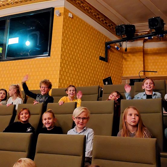Kinder sitzen in Reihen in einem Theater, einige mit erhobenen Händen, lächeln, tragen Brillen und einige mit Pferdeschwänzen. Das Theater hat gelbe Wände, eine große Leinwand und an der Decke montierte Lautsprecher.