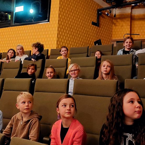 Mehrere Kinder sitzen in einem Auditorium mit gelben sechseckigen Wänden und Deckenleuchten. Sie sehen sich einen Film oder eine Präsentation an.