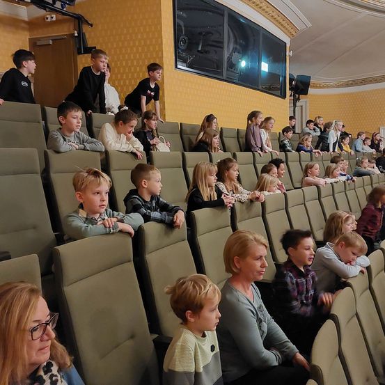 Ein Auditorium gefüllt mit Kindern und Erwachsenen, einige stehend und andere auf grünen Stühlen sitzend, die eine Leinwand betrachten. Einige Kinder schauen zur Kamera, während andere in der Show engagiert sind.