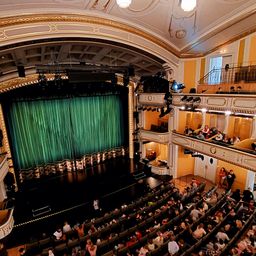 Ein Publikum sitzt in einem Theater und sieht eine Aufführung auf der Bühne. Die Bühne hat einen großen grünen Vorhang, und das Theater ist mit Gold und Gelb dekoriert.
