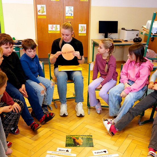 Eine Gruppe von Kindern und einer Frau sitzen im Kreis. Die Frau hält ein kreisförmiges Objekt mit Text. Kinder schauen die Frau an. Karten mit Text liegen auf dem Boden.