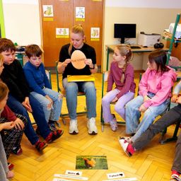 Eine Gruppe von Kindern und einer Frau sitzen im Kreis. Die Frau hält ein kreisförmiges Objekt mit Text. Kinder schauen die Frau an. Karten mit Text liegen auf dem Boden.
