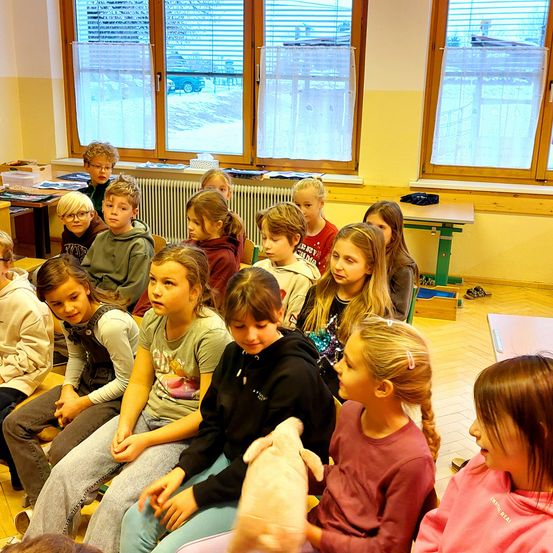 In einem Klassenzimmer sitzen mehrere Kinder aufmerksam vor einer Lehrerin, mit Tischen und Fenstern dahinter.