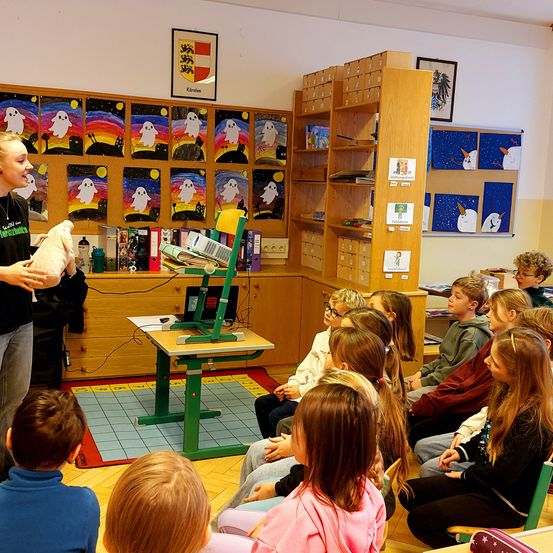 Eine Frau, die ein Kuscheltier hält, steht vor einem Klassenzimmer voller Kinder, mit bunten Gemälden an der Wand, einem Projektor auf einem Schreibtisch und Regalen voller Bücher.