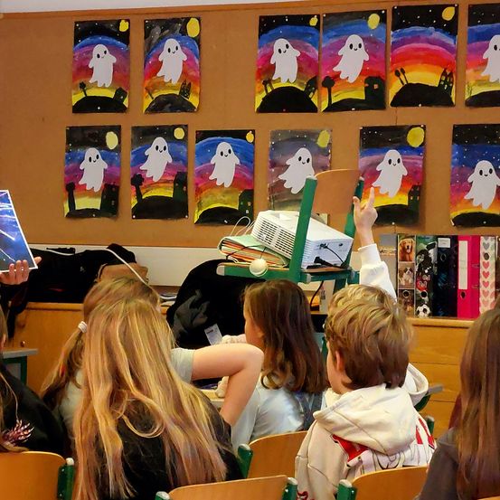 Ein Klassenzimmer mit farbenfrohen Geistergemälden an der Wand. Schüler sitzen in Stühlen, einer hebt die Hand. Ein Beamer steht auf einem Ständer, und eine Person hält ein Buch.