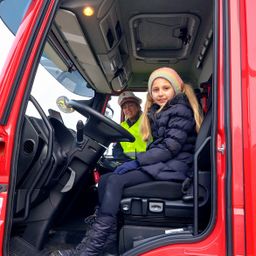 Ein junges Mädchen sitzt lächelnd auf dem Fahrersitz eines roten Feuerwehrautos. Eine Frau in einer Warnweste steht hinter ihr und hält das Lenkrad.