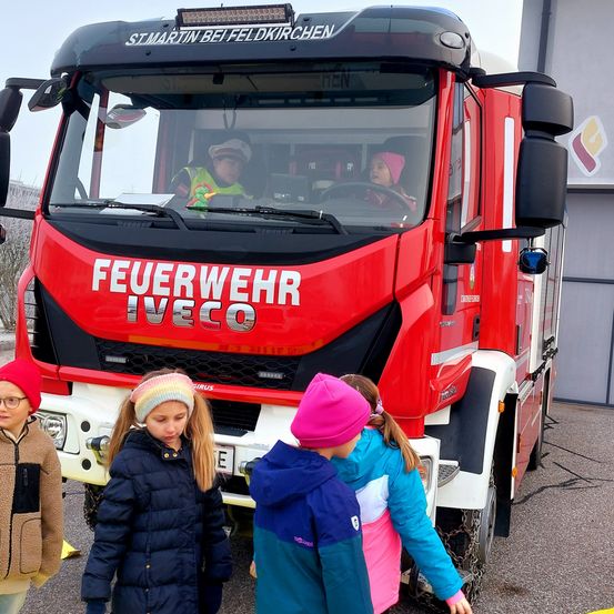 Ein Feuerwehrfahrzeug mit der Aufschrift 'Feuerwehr IVECO' auf der Front ist geparkt. Ein Mann fährt das Fahrzeug. Vier Kinder stehen vor dem Fahrzeug.