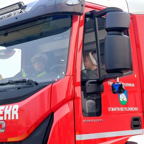 Ein roter Feuerwehrwagen mit Fahrer und Beifahrer im Inneren. Der Wagen ist mit Eurocargo beschriftet und hat ein Logo mit Text auf der Tür.
