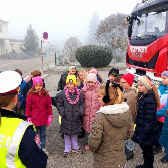 Eine Gruppe von Kindern in Wintermänteln steht vor einem roten Feuerwehrauto, mit einem Polizisten in der Nähe, an einem nebligen Tag.