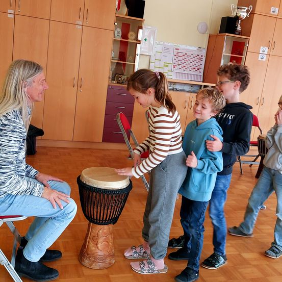 Ein älterer Mann bringt einem jungen Mädchen und zwei Jungen das Schlagzeugspielen in einem Raum mit Holzschränken und ausgestellten Trophäen bei.