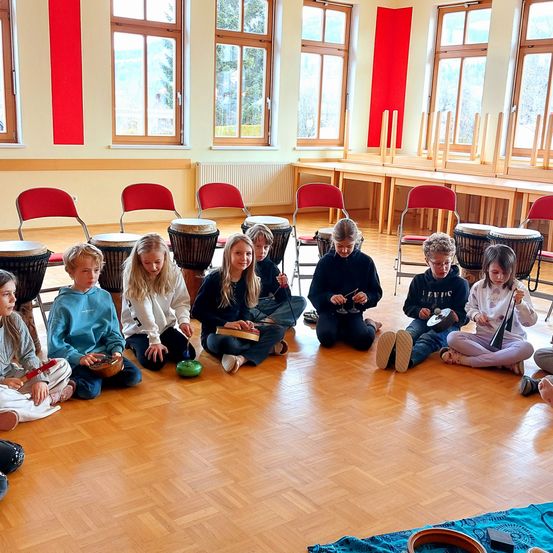 Eine Gruppe von Kindern sitzt im Kreis und hält verschiedene Musikinstrumente wie Trommeln und Becken in den Händen. Sie schauen zu einem Erwachsenen, der sie unterrichtet. Hinter ihnen stehen mehrere rote Stühle und Tische. Der Raum hat Holzböden und weiße Wände mit roten Akzenten.