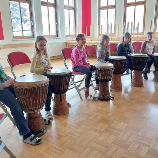 Kinder sitzen in einem Raum und spielen Trommeln. Sie sind alle lässig gekleidet. Hinter ihnen sind große Fenster und ein Heizkörper zu sehen.