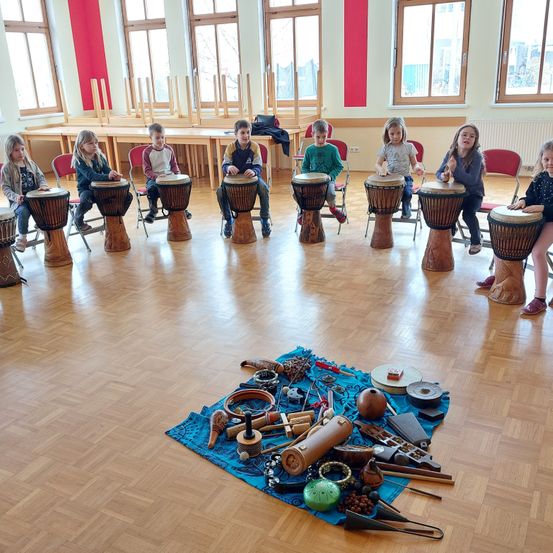 Eine Gruppe von Kindern sitzt in Stühlen in einem Kreis und spielt Trommeln, mit verschiedenen Musikinstrumenten auf einem blauen Tuch in der Mitte.