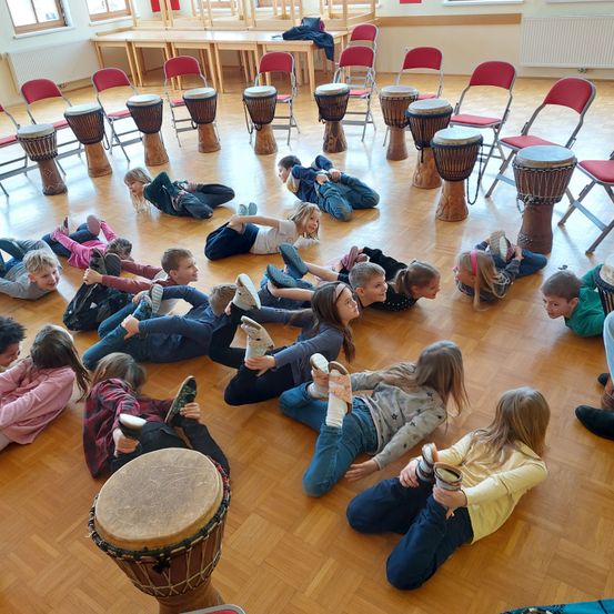 Eine Gruppe von Kindern liegt auf dem Boden um Trommeln herum und übt ihr Trommeln. Der Raum hat Holzböden und weiße Wände.