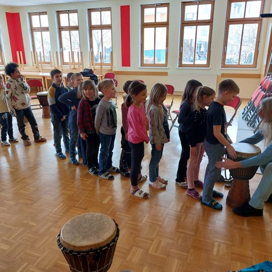 Eine Gruppe von Kindern steht in einer Reihe in einem Raum mit Holzboden. Es gibt Trommeln auf dem Boden und eine Frau, die eine spielt. Der Raum hat große Fenster und rote Wände.
