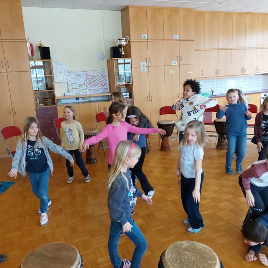 Eine Gruppe von Kindern in einem Klassenzimmer tanzt um Trommeln, während einige auf Stühlen sitzen. Hinter ihnen befinden sich Holzschränke und Trophäen sind an der Wand ausgestellt.