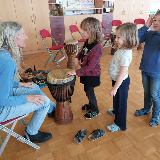 Ein älterer Mann mit langen Haaren sitzt auf einem Stuhl und spielt Trommel. Zwei junge Mädchen stehen vor ihm, eines hält eine Trommel. Ein Junge steht hinter ihnen. Sie sind in einem Raum mit Holzböden und roten Stühlen.