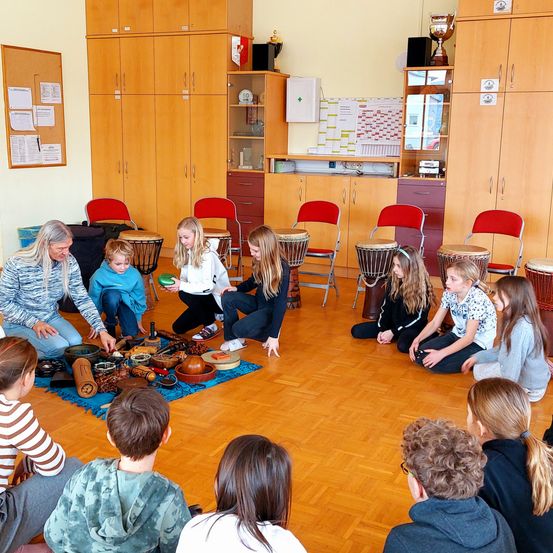 Mehrere Kinder und eine erwachsene Frau versammeln sich in einem Klassenzimmer mit Trommeln und anderen Instrumenten. Das Klassenzimmer hat Holzschränke und rote Stühle.