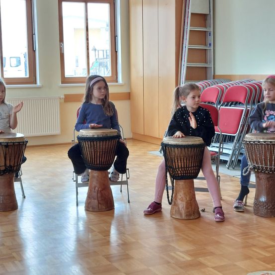 Vier Mädchen sitzen auf Stühlen und spielen jeweils ein Djembe. Der Raum hat Holzböden und eine weiße Wand mit Fenstern. Dahinter sind eine Reihe roter Stühle und eine Leiter zu sehen.