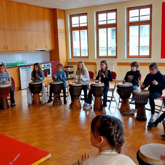 Eine Gruppe von Kindern spielt in einem Raum mit Holzboden und großen Fenstern Trommeln. Jedes Kind sitzt auf einem Stuhl und spielt eine Trommel.