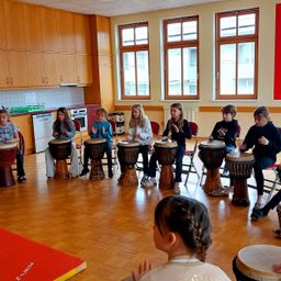 Eine Gruppe von Kindern spielt in einem Raum mit Holzboden und großen Fenstern Trommeln. Jedes Kind sitzt auf einem Stuhl und spielt eine Trommel.