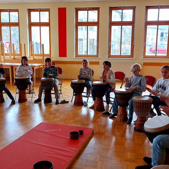 Mehrere Kinder sitzen in einem Kreis auf Stühlen um Trommeln herum und üben das Spielen. Sie befinden sich in einem Raum mit großen Fenstern und einem roten Bodenteppich.