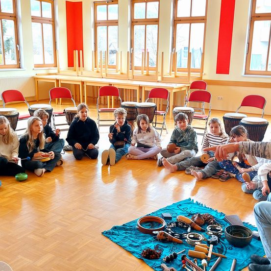 Eine Gruppe von Kindern sitzt auf dem Boden und spielt Trommeln und andere Musikinstrumente. Sie befinden sich in einem Raum mit Holzböden und Fenstern.