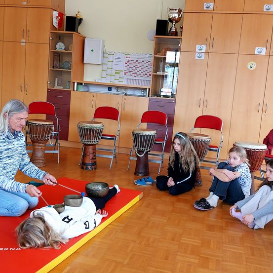 Ein erwachsener Mann spielt Trommeln, während ein Mädchen auf einer roten Matte liegt und drei Mädchen auf Stühlen in einem Klassenzimmer sitzen.