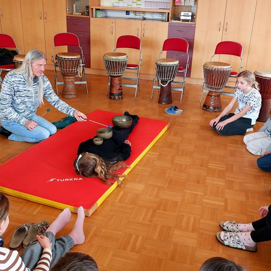 Ein älterer Mann unterrichtet Kinder in einem Raum mit Holzböden und Stühlen. Die Kinder sitzen herum, und eines liegt auf einer roten Matte mit Schüsseln, während der Lehrer ein Schlagzeug spielt.