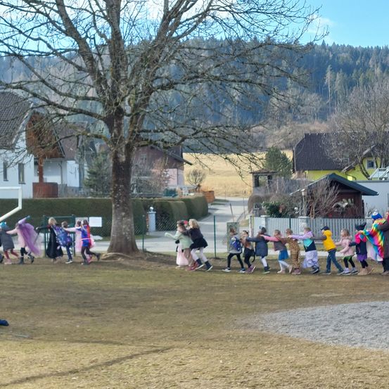 Eine Gruppe von Kindern in Kostümen bildet einen Kreis auf einem Grasfeld vor einem Haus mit einem Baum, unter einem klaren Himmel mit Bergen im Hintergrund.
