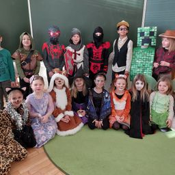 Eine Gruppe von Kindern in Kostümen posiert für ein Foto im Klassenzimmer. Sie sind als verschiedene Charaktere verkleidet, darunter ein Ritter, Spiderman und eine Hexe.