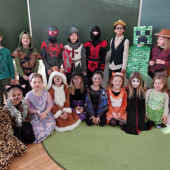 Eine Gruppe von Kindern in Kostümen posiert für ein Foto im Klassenzimmer. Sie sind als verschiedene Charaktere verkleidet, darunter ein Ritter, Spiderman und eine Hexe.