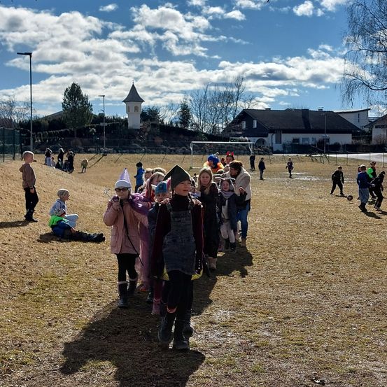 Eine Gruppe von Kindern, einige in Kostümen, geht auf einem Feld unter einem bewölkten Himmel. In der Nähe stehen oder sitzen Menschen an einem Hang. Im Hintergrund befinden sich Häuser, ein Turm und ein Fußballtor.