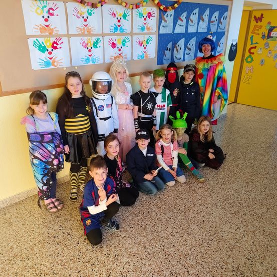 Eine Gruppe von Kindern in verschiedenen Kostümen posiert für ein Foto in einem Klassenzimmer. Sie sitzen und stehen vor einer Wand, die mit Handabdrücken und Zeichnungen verziert ist.