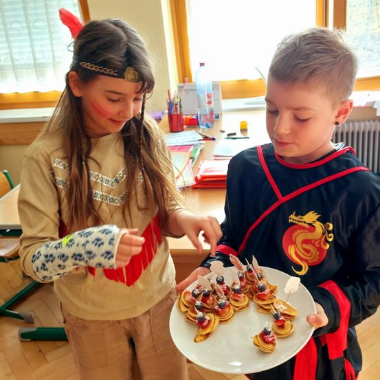 Ein junges Mädchen mit Gesichtsbemalung und Kopfband schaut einen Jungen mit Drachenhemd an, der einen Teller mit Essen hält.