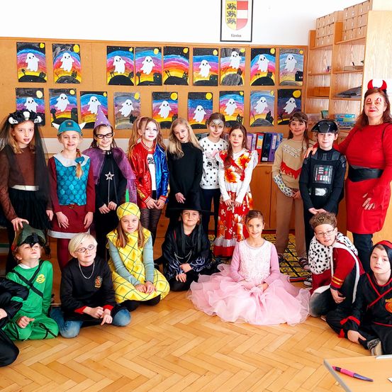 Ein Klassenzimmer voller Kinder in verschiedenen Kostümen, einige stehend und andere sitzend, alle posieren für ein Gruppenfoto mit Halloween-Dekorationen an der Wand.