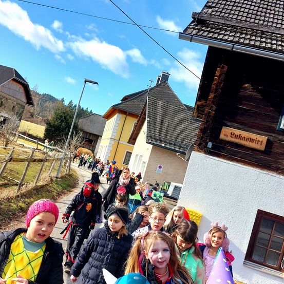 Eine Gruppe von Kindern in verschiedenen Kostümen geht eine Straße entlang, die von Häusern gesäumt ist, eines mit einem Schild, das 'Gukaschmid' liest. Im Hintergrund sind ein Feld und ein Hügel unter einem teilweise bewölkten Himmel sichtbar.