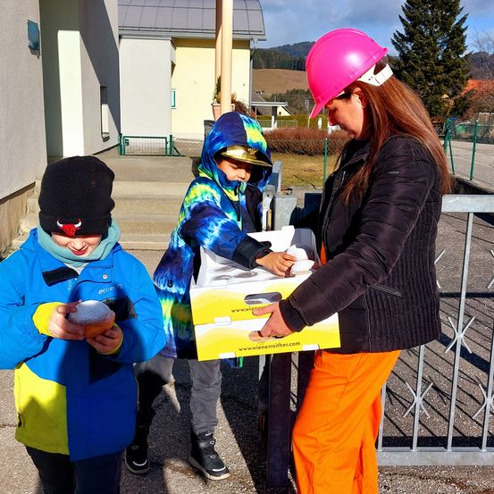 Eine Frau in einem rosa Helm reicht zwei Kindern Döngel aus. Ein Kind hält einen Donut und das andere einen Beutel. Sie stehen auf einem Bürgersteig, mit einem Gebäude, Metallzaun und Bergen im Hintergrund.