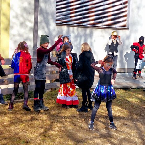 Eine Gruppe von Kindern in verschiedenen Kostümen steht vor einem Gebäude mit weißer Wand und Metalljalousien. Einige schauen in Richtung des Gebäudes, andere nach unten.