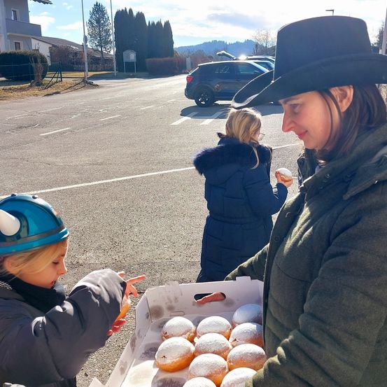 Eine Frau in einem Cowboyhut hält eine Schachtel mit Donuts, während ein Kind in einem Helm darauf zeigt. Eine weitere Frau und ein Mädchen sind im Hintergrund, und ein Auto ist in der Nähe geparkt.