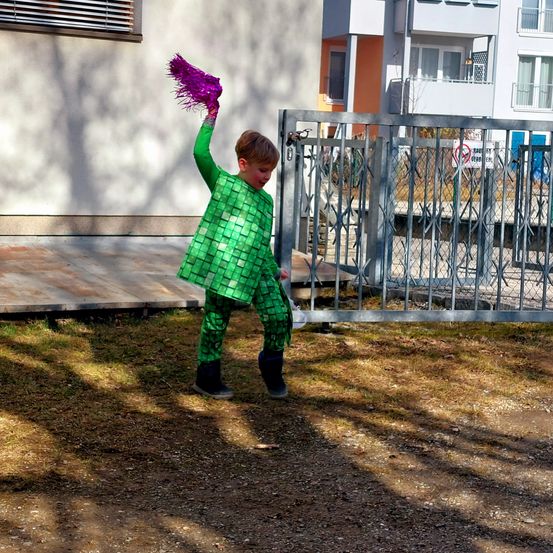 Ein junger Junge in einem grünen Pailletten-Outfit hält eine lila Feder in der Luft. Er steht auf einer Rasenfläche vor einem Gebäude mit einem Metalltor.